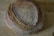 A round loaf of artisan bread with a crispy, textured crust and a deep brown color rests on a wooden surface. The bread's surface features a pattern of concentric cracks and lines, highlighting its rustic quality and artisanal craftsmanship.