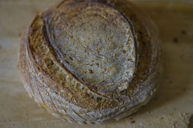 A round loaf of artisan bread with a crispy, textured crust and a deep brown color rests on a wooden surface. The bread's surface features a pattern of concentric cracks and lines, highlighting its rustic quality and artisanal craftsmanship.