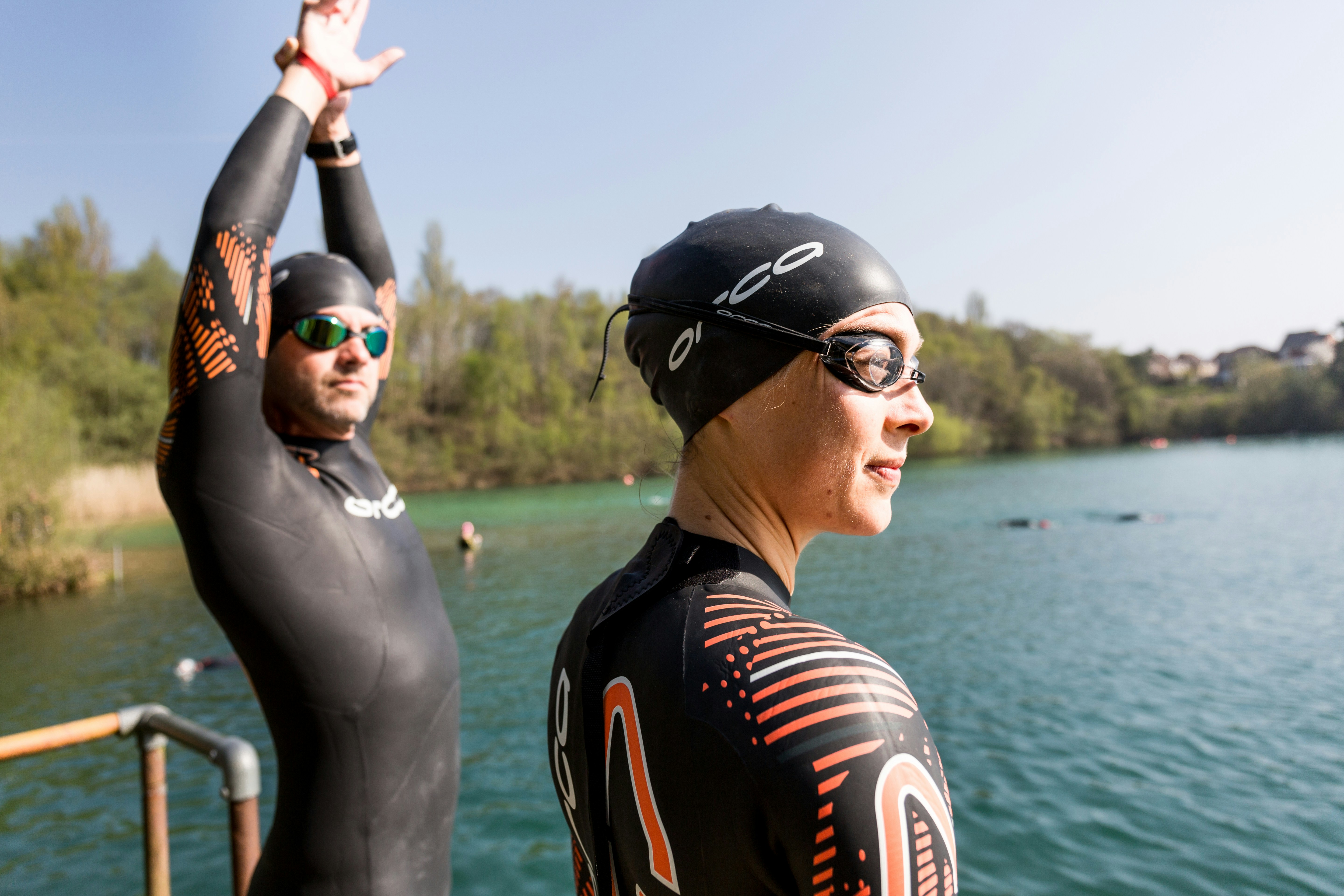 Two people wearing wetsuits and swim caps stand on the edge of a body of water, preparing for open-water swimming. One person is stretching with their arms raised, while the other looks out across the water. Trees and foliage line the opposite bank under a clear sky.