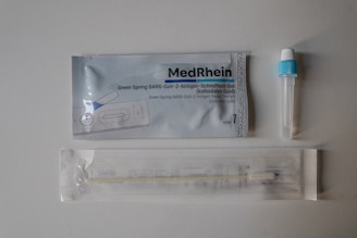 The image displays components of a COVID-19 rapid antigen test kit. There is a sealed packet with branding and instructions, a small vial with a blue cap, and a packaged swab. The items are arranged on a light gray surface.
