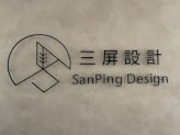 A minimalist logo with a circular geometric design and a stylized tree inside a partial circle. Accompanied by text in both Chinese characters and the English words 'SanPing Design' on a textured gray background.