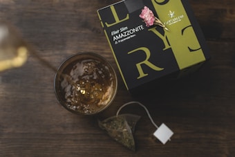 A glass filled with a golden liquid sits next to a box of tea labeled 'Elixir Slim AMAZZONITE.' The box is adorned with large letters in a modern font and features a decorative pink flower on top. A tea bag with visible dried leaves is placed nearby on a wooden surface.