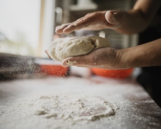 Hands are holding a piece of dough over a flour-dusted surface in a warm, softly lit kitchen setting. A sprinkle of flour is visible in the air, evoking a sense of motion and preparation.