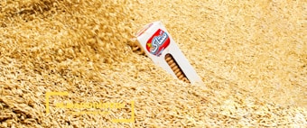 A box of wheat biscuits is partially buried in a large pile of wheat grains. The image foreground and background are filled with the light brown and golden hues of the grains, creating a textured look. The biscuit box has a clear front display showing vertical stacks of the biscuits and a logo in red and blue.