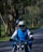 A person wearing a blue and white jacket and a helmet rides a motorcycle on a tree-lined road. The scene is filled with greenery, suggesting a rural or forest setting. A second person appears seated behind, also wearing a helmet, holding a phone.
