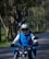 A person wearing a blue and white jacket and a helmet rides a motorcycle on a tree-lined road. The scene is filled with greenery, suggesting a rural or forest setting. A second person appears seated behind, also wearing a helmet, holding a phone.