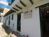 A quaint white building with green window and door frames, featuring a sign that reads 'la moraleja - villa de leyva.' The building has a rustic stone path in front, and pots with plants are visible. Bright string lights are hanging near the entrance.
