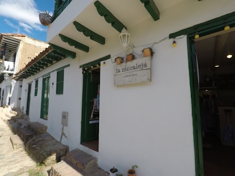 A quaint white building with green window and door frames, featuring a sign that reads 'la moraleja - villa de leyva.' The building has a rustic stone path in front, and pots with plants are visible. Bright string lights are hanging near the entrance.