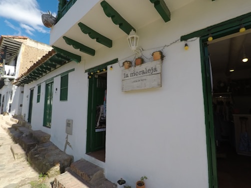 A quaint white building with green window and door frames, featuring a sign that reads 'la moraleja - villa de leyva.' The building has a rustic stone path in front, and pots with plants are visible. Bright string lights are hanging near the entrance.