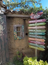 A rustic wall made of adobe, topped with a clay-tiled roof, features a small, weathered window adorned with colorful floral patterns. Next to the window is a vibrant wooden signpost with multiple arrows pointing in different directions, each labeled with menu items such as 'Mojarra Asada', 'Chuleta de Cordero', and 'Salmon a la Brasa'. Lush green foliage and greenery surround the base and part of the wall, adding to the quaint and inviting setting.