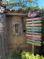 A rustic wall made of adobe, topped with a clay-tiled roof, features a small, weathered window adorned with colorful floral patterns. Next to the window is a vibrant wooden signpost with multiple arrows pointing in different directions, each labeled with menu items such as 'Mojarra Asada', 'Chuleta de Cordero', and 'Salmon a la Brasa'. Lush green foliage and greenery surround the base and part of the wall, adding to the quaint and inviting setting.