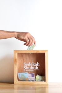A hand is placing a banknote into a wooden donation box, which contains various other banknotes and coins. The box has 'Sedekah Shubuh' written on it along with a social media handle.