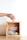 A hand is placing a banknote into a wooden donation box, which contains various other banknotes and coins. The box has 'Sedekah Shubuh' written on it along with a social media handle.