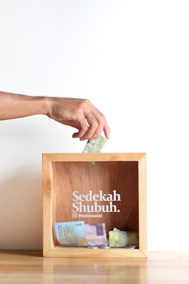 A hand is placing a banknote into a wooden donation box, which contains various other banknotes and coins. The box has 'Sedekah Shubuh' written on it along with a social media handle.