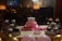 A festive birthday setting features a cake with a pink frosting base and a small top layer decorated with polka dots. Surrounding the cake are chocolate cupcakes, each topped with whipped cream and colorful candies. A pink 'Birthday Girl' sash is displayed in front of the cake along with a small crown. The table is adorned with pink and gold balloons, and the background shows a dimly lit room with decorative wallpaper.