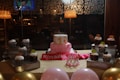 A festive birthday setting features a cake with a pink frosting base and a small top layer decorated with polka dots. Surrounding the cake are chocolate cupcakes, each topped with whipped cream and colorful candies. A pink 'Birthday Girl' sash is displayed in front of the cake along with a small crown. The table is adorned with pink and gold balloons, and the background shows a dimly lit room with decorative wallpaper.