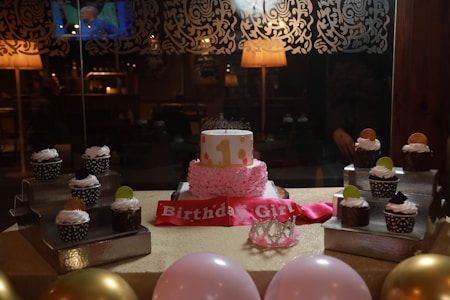 A festive birthday setting features a cake with a pink frosting base and a small top layer decorated with polka dots. Surrounding the cake are chocolate cupcakes, each topped with whipped cream and colorful candies. A pink 'Birthday Girl' sash is displayed in front of the cake along with a small crown. The table is adorned with pink and gold balloons, and the background shows a dimly lit room with decorative wallpaper.