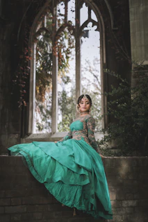 A regal model draped in an emerald green silk velvet ensemble, standing against a backdrop of Mughal-inspired arches bathed in soft champagne gold light.