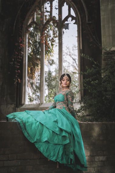 A regal model draped in an emerald green silk velvet ensemble, standing against a backdrop of Mughal-inspired arches bathed in soft champagne gold light.