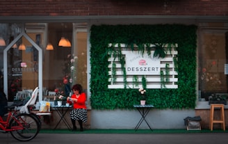 A cozy cafe storefront with a sign reading 'Desszert' set against a vibrant wall of green foliage. A woman in a red top and polka dot skirt sits at a small table, enjoying some refreshments. A red bicycle with a child seat is parked nearby, adding to the inviting urban scene.