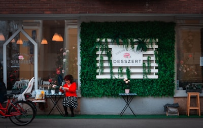 A cozy cafe storefront with a sign reading 'Desszert' set against a vibrant wall of green foliage. A woman in a red top and polka dot skirt sits at a small table, enjoying some refreshments. A red bicycle with a child seat is parked nearby, adding to the inviting urban scene.