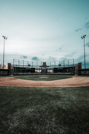 An empty baseball field with a dirt diamond, surrounded by grass. The sky is overcast, and there are tall floodlights on either side of the field. The backstop and a small building are visible in the distance.