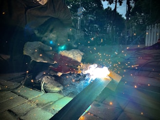 A person wearing work gloves is engaged in welding on a metal surface. Sparks and bright light are emitted from the welding area, creating a dramatic contrast against the darker surroundings. The ground is paved with bricks, and there are some tools and equipment scattered around.