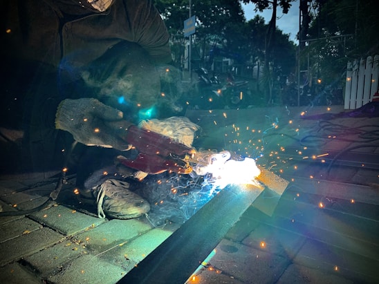 A person wearing work gloves is engaged in welding on a metal surface. Sparks and bright light are emitted from the welding area, creating a dramatic contrast against the darker surroundings. The ground is paved with bricks, and there are some tools and equipment scattered around.