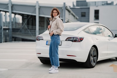 A person stands in a parking lot next to a white car, wearing a beige jacket and blue jeans. The background features a modern, industrial-looking multi-story structure.