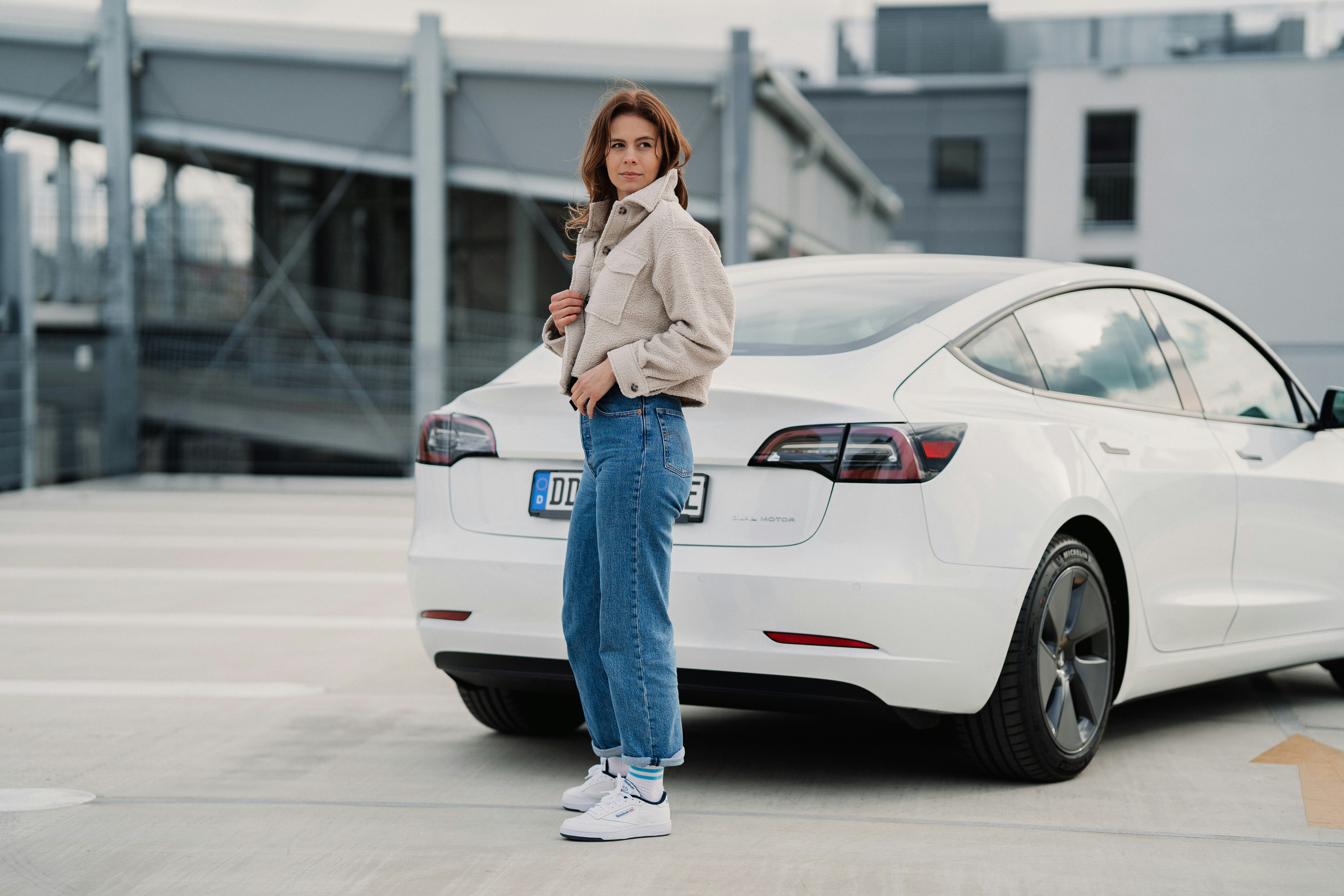 woman in white jacket and blue denim jeans standing beside white car during daytime