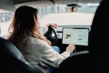 A person is sitting in the driver's seat of a modern car, interacting with a large touch screen display on the dashboard. The car appears sleek and technologically advanced, with a focus on the digital interface. The person's attention is on the screen, suggesting involvement with navigation or vehicle settings.