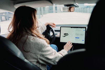 A person is sitting in the driver's seat of a modern car, interacting with a large touch screen display on the dashboard. The car appears sleek and technologically advanced, with a focus on the digital interface. The person's attention is on the screen, suggesting involvement with navigation or vehicle settings.