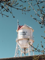 A white water tower with the text 'Burns & Wilcox' is prominently displayed against a clear blue sky. The tower is surrounded by tree branches with budding leaves, and an American flag is mounted on the tower.