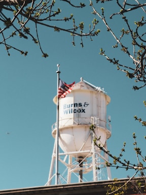 A white water tower with the text 'Burns & Wilcox' is prominently displayed against a clear blue sky. The tower is surrounded by tree branches with budding leaves, and an American flag is mounted on the tower.