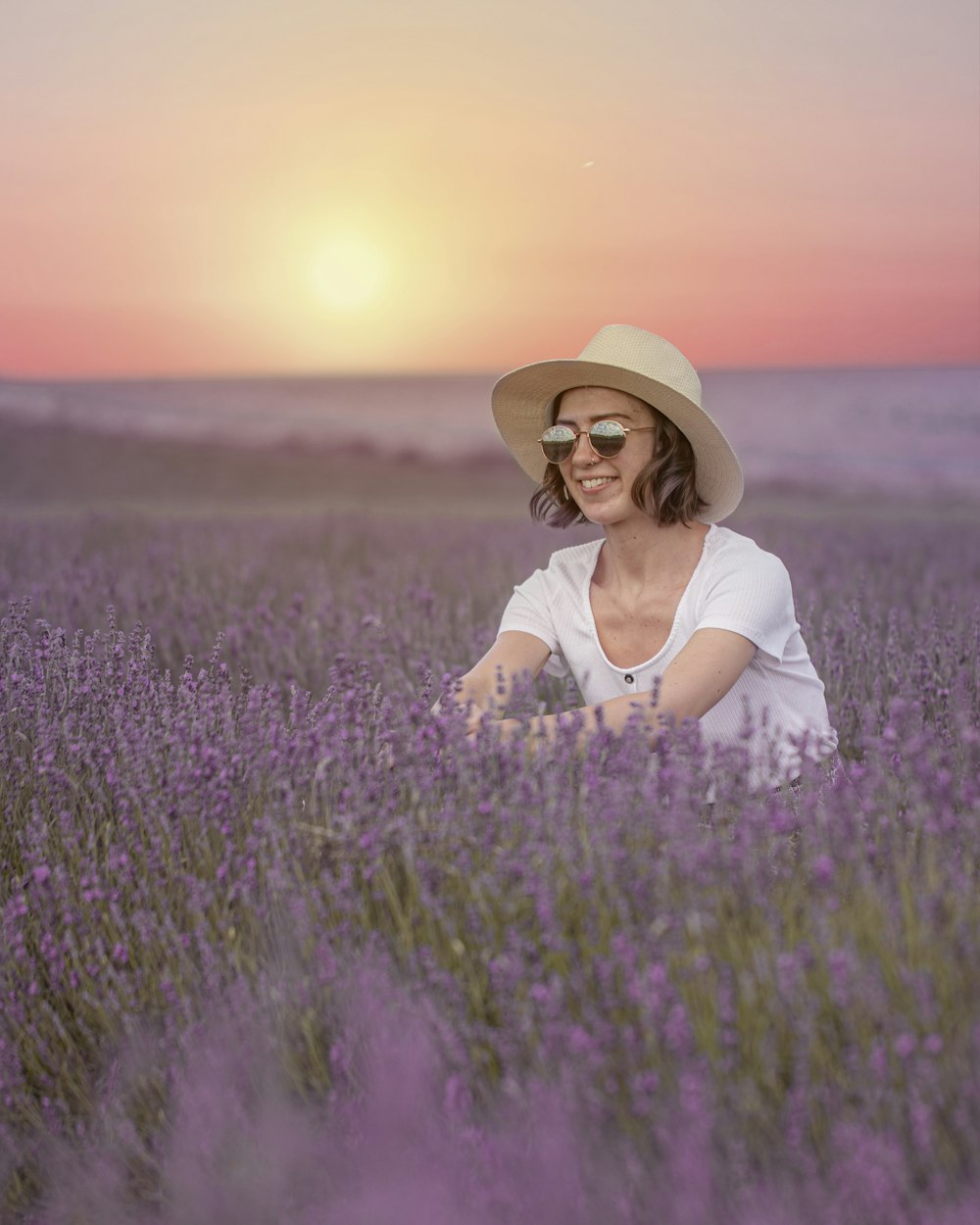 Donna in camicia bianca e cappello marrone che si siede sul campo di fiori viola durante il giorno