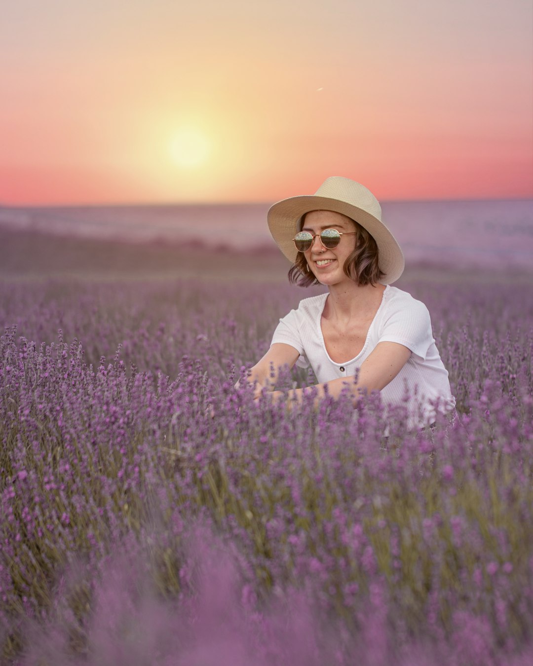 woman in white shirt and brown hat sitting on purple flower field during daytime