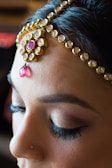 A close-up of a woman's face featuring intricately designed gold jewelry on the forehead with embedded stones, including a prominent pink stone with a teardrop shape. The woman has closed eyes with well-groomed eyebrows and long eyelashes. There is subtle makeup applied, accentuating the natural features.