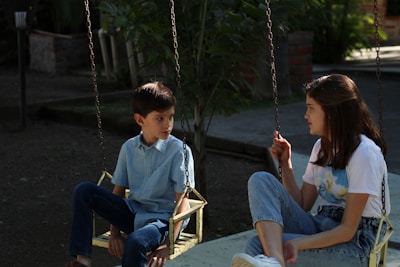 A young boy and a girl are sitting on separate swings. They appear to be engaged in a conversation, with the girl attentively listening. The surroundings are lush with greenery, and the area looks like a park or garden setting. Both individuals are wearing casual attire - the boy in a light blue button-up shirt and jeans, and the girl in a graphic t-shirt and jeans.