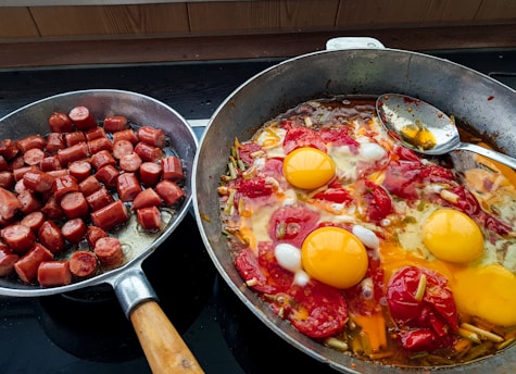 The image displays two frying pans on a stove. The left pan contains chopped sausages, cooked until they are browned. The right pan holds a mix of vegetables, tomatoes, and three cracked eggs, all cooking together with a spoon resting inside.