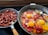 The image displays two frying pans on a stove. The left pan contains chopped sausages, cooked until they are browned. The right pan holds a mix of vegetables, tomatoes, and three cracked eggs, all cooking together with a spoon resting inside.