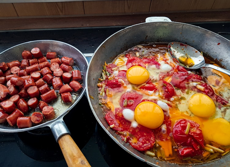 The image displays two frying pans on a stove. The left pan contains chopped sausages, cooked until they are browned. The right pan holds a mix of vegetables, tomatoes, and three cracked eggs, all cooking together with a spoon resting inside.