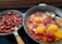 The image displays two frying pans on a stove. The left pan contains chopped sausages, cooked until they are browned. The right pan holds a mix of vegetables, tomatoes, and three cracked eggs, all cooking together with a spoon resting inside.