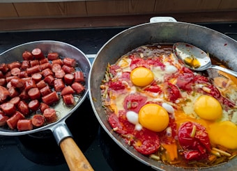 The image displays two frying pans on a stove. The left pan contains chopped sausages, cooked until they are browned. The right pan holds a mix of vegetables, tomatoes, and three cracked eggs, all cooking together with a spoon resting inside.