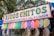 A colorful street vendor sign displaying a variety of candy or snack items, each wrapped in clear packaging and clipped to a string. The sign above reads 'Ricos Chitos, Preparados con Chile, Sal y Limon' in bold letters. The background features green trees, suggesting an outdoor setting.