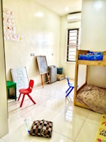 A small, bright room with a tiled floor, containing children's furniture such as colorful plastic chairs, a small table, and a bunk bed. The walls have educational materials like alphabet cutouts and whiteboards with scribbles. Various items, including a chalkboard, cushion, and water bottle, are scattered around.