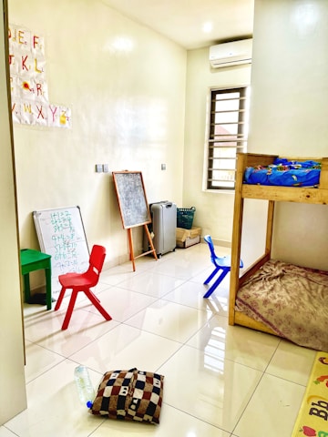 A small, bright room with a tiled floor, containing children's furniture such as colorful plastic chairs, a small table, and a bunk bed. The walls have educational materials like alphabet cutouts and whiteboards with scribbles. Various items, including a chalkboard, cushion, and water bottle, are scattered around.