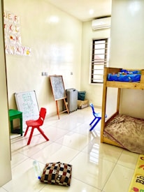 A small, bright room with a tiled floor, containing children's furniture such as colorful plastic chairs, a small table, and a bunk bed. The walls have educational materials like alphabet cutouts and whiteboards with scribbles. Various items, including a chalkboard, cushion, and water bottle, are scattered around.