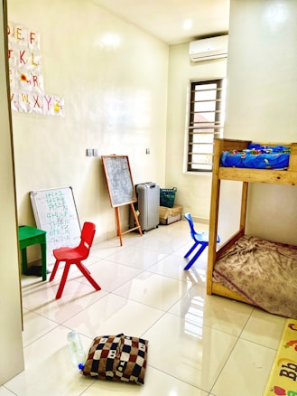 A small, bright room with a tiled floor, containing children's furniture such as colorful plastic chairs, a small table, and a bunk bed. The walls have educational materials like alphabet cutouts and whiteboards with scribbles. Various items, including a chalkboard, cushion, and water bottle, are scattered around.