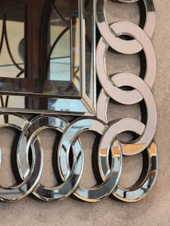 A decorative mirror with an intricate frame composed of interlocking circular metallic rings. The surface has a reflective quality, and the background appears to be a textured beige wall. The decorative elements display a sophisticated design.