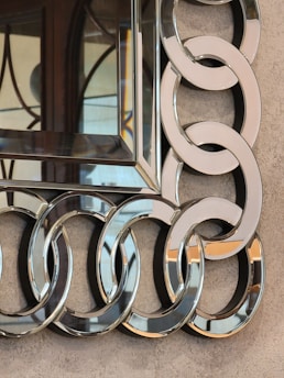 A decorative mirror with an intricate frame composed of interlocking circular metallic rings. The surface has a reflective quality, and the background appears to be a textured beige wall. The decorative elements display a sophisticated design.